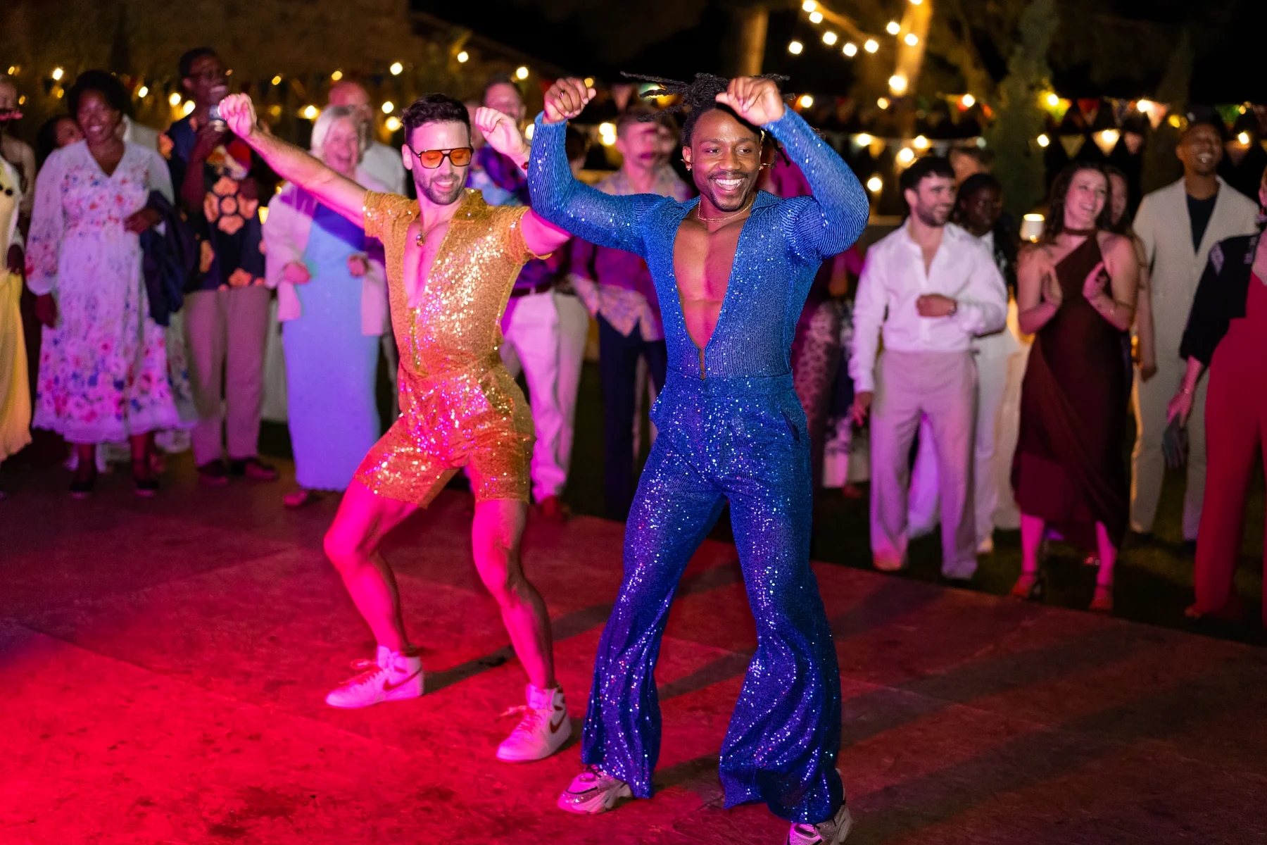 Two grooms in sequined disco-style jumpsuits dance joyfully at an outdoor reception at Conti di San Bonifacio in Tuscany.