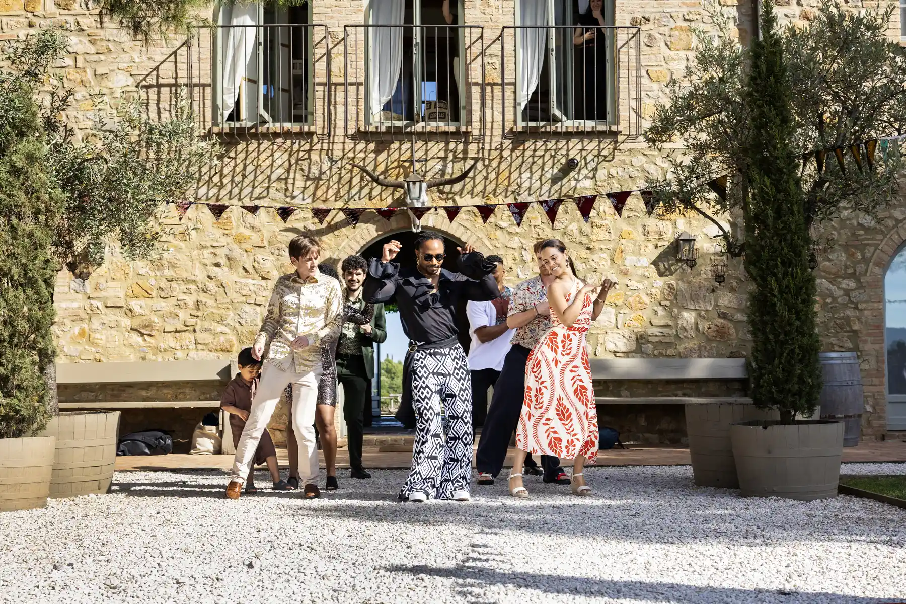 Wedding guests and grooms dance outdoors at Conti di San Bonifacio Wine Resort in Tuscany, Italy, on a sun-drenched gravel courtyard.