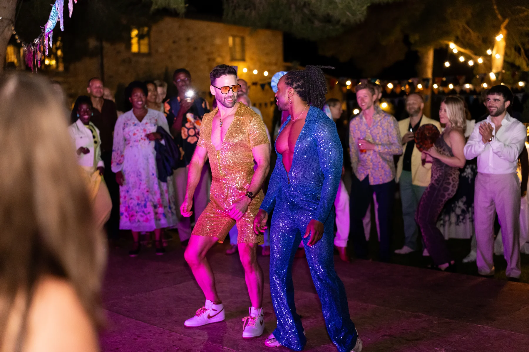 Two grooms in sequined jumpsuits dance together on an outdoor dance floor at Conti di San Bonifacio Wine Resort in Tuscany.